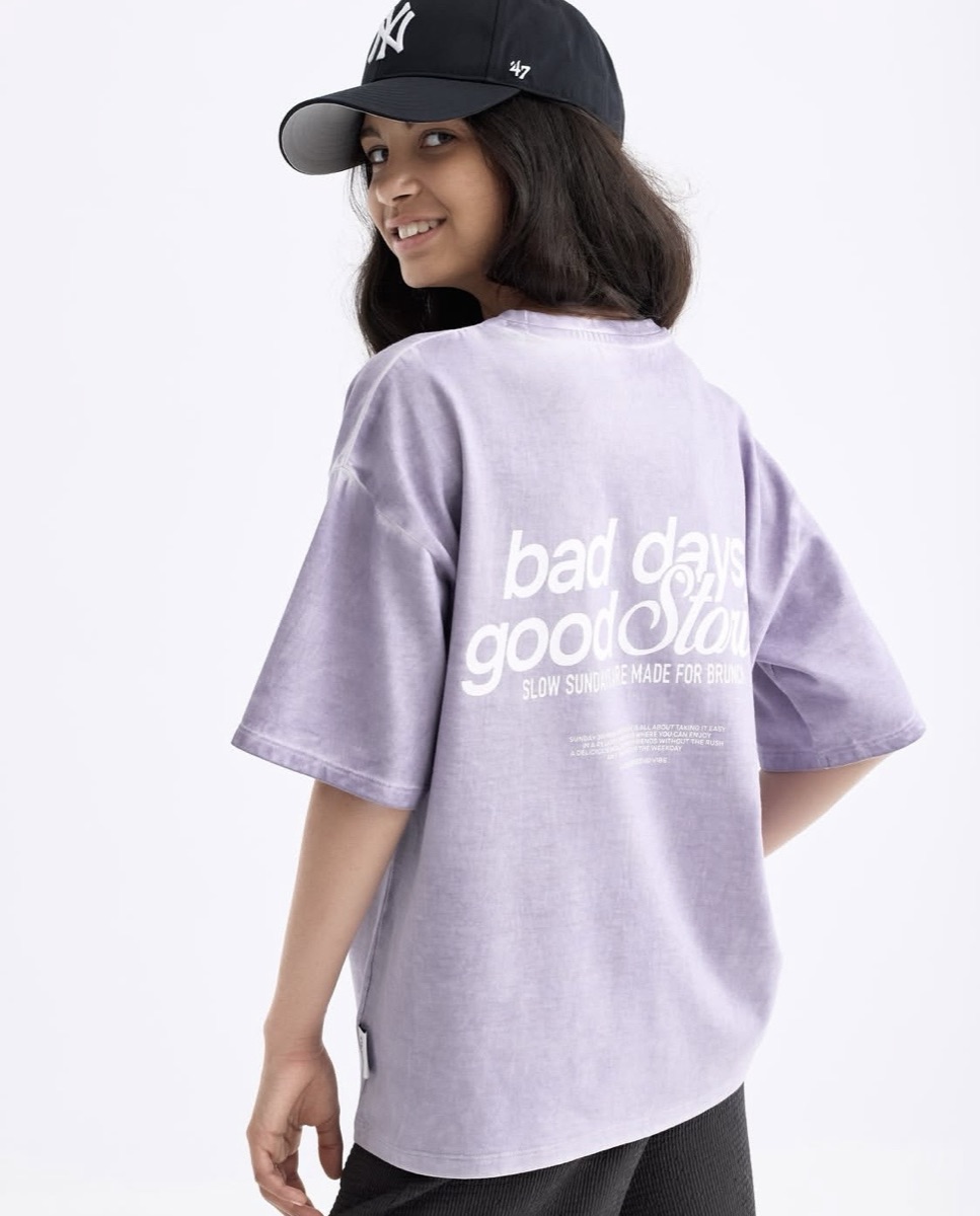 A girl in an oversized garment-washed lilac T-shirt with a large white typographic back print